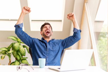 Neşeli iş adamı, evinde masa başında oturup dizüstü bilgisayarla çalışırken kollarını havaya kaldırarak kutlama yapıyor. Merkez Ofis.
