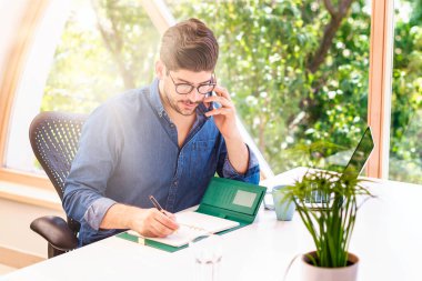 Mutlu bir iş adamı iş görüşmesi yaparken ve dizüstü bilgisayarının arkasında oturup evde çalışırken not defterine bir şeyler yazarken. Merkez Ofis. 