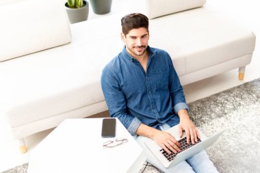 Mutlu adamın sıradan kıyafetler giydiği ve evde çalışırken dizüstü bilgisayar kullandığı yüksek açılı bir çekim. Merkez Ofis.