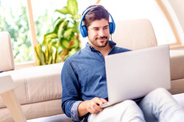 Kanepede oturup dizüstü bilgisayar kullanırken, kulaklıklı, müzik dinleyen rahat bir adamın fotoğrafı..