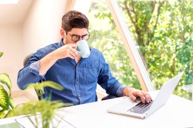 Ofis masasında dizüstü bilgisayarının arkasında oturan ve evden çay içen bir iş adamı. Merkez Ofis.