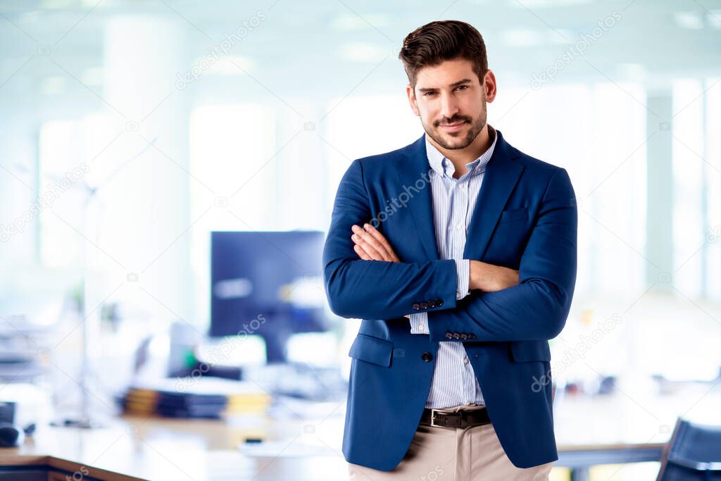 Portrait of handsome businessman wearing shirt and blazer while standing with arms crossed in the office. 