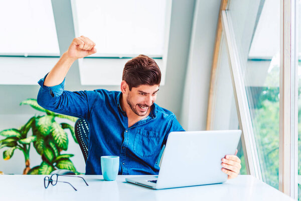 Cheerful businessman celebrating with his arm raised in the air while sitting at a desk in his home office and working on a laptop. Home office.