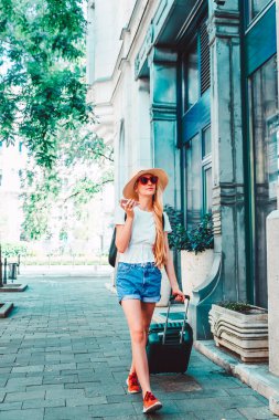 Sokakta bavuluyla yürürken ve cep telefonuyla konuşurken hasır şapka ve sırt çantası takan genç bir kadın turistin tam boy görüntüsü.. 