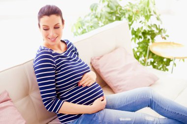 Güzel hamile bir kadının bebek çanının üstünde evinde koltukta dinlenirken hamile olduğunu gösteren yüksek açılı bir fotoğraf..