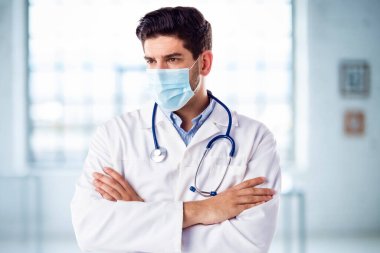 Hastanenin fuayesinde kolları çapraz dururken maske takan erkek doktorun portresi.