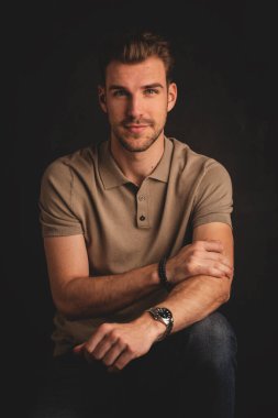 Tecrit edilmiş arka planda oturan yakışıklı bir gencin stüdyo fotoğrafı. Polo tişört giyen ve kameraya bakan kendinden emin bir erkek. Uzayı kopyala İzole edilmiş koyu arkaplan.