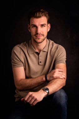 Tecrit edilmiş arka planda oturan yakışıklı bir gencin stüdyo fotoğrafı. Polo tişört giyen ve kameraya bakan kendinden emin bir erkek. Uzayı kopyala İzole edilmiş koyu arkaplan.