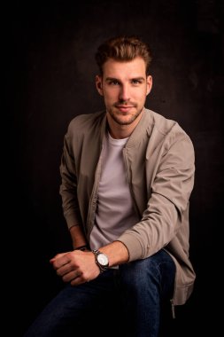 Tecrit edilmiş arka planda oturan yakışıklı bir gencin stüdyo fotoğrafı. Tişört ve ceket giyen ve kameraya bakan kendinden emin bir erkek. Uzayı kopyala İzole edilmiş koyu arkaplan.