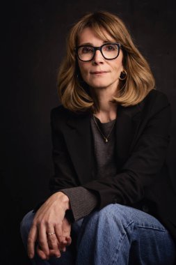 Sarışın kadın, izole edilmiş siyah arka planda güvenle oturuyor ve kameraya bakıyor. Ceket ve kot pantolon giyen orta yaşlı bir kadın. Boşluğu kopyala. 