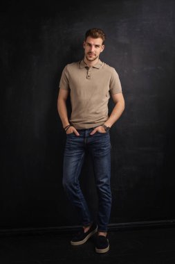 Tecrit edilmiş arka planda duran yakışıklı bir gencin stüdyo fotoğrafı. Polo tişört giyen ve kameraya bakan kendinden emin bir erkek. Uzayı kopyala İzole edilmiş koyu bir arka plan. Tam uzunluk.