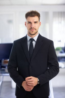 Modern bir ofiste tek başına dururken gülümseyen kendinden emin genç bir iş adamının portresi. Takım elbise ve kravat takan genç bir girişimci. Yakın çekim..