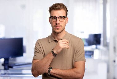 Modern bir ofiste tek başına dururken gülümseyen kendinden emin genç bir iş adamının vesikalığı. Gündelik kıyafetler giyen genç bir girişimci. Yakın çekim..