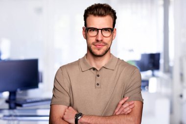 Modern bir ofiste tek başına dururken gülümseyen kendinden emin genç bir iş adamının vesikalığı. Gündelik kıyafetler giyen genç bir girişimci. Yakın çekim..