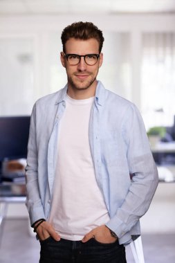Modern bir ofiste tek başına dururken gülümseyen kendinden emin genç bir iş adamının portresi. Gündelik kıyafetler giyen genç bir girişimci. Yakın çekim..
