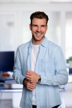 Modern bir ofiste tek başına dururken gülümseyen kendinden emin genç bir iş adamının portresi. Gündelik kıyafetler giyen genç bir girişimci. Yakın çekim..