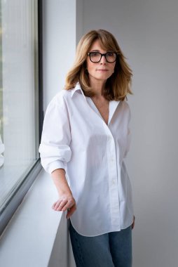 Kendine güvenen, sarı saçlı kadın pencerenin önünde duruyor. Orta yaşlı kadın beyaz gömlek ve gözlük takıyor.. 