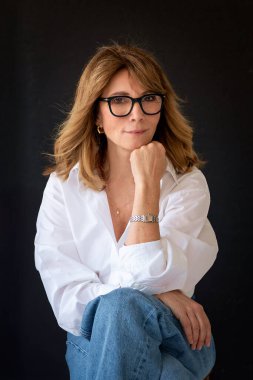 Sarışın kadın, izole edilmiş siyah arka planda güvenle oturuyor ve kameraya bakıyor. Beyaz gömlek ve kot pantolon giyen orta yaşlı bir kadın. Boşluğu kopyala. 