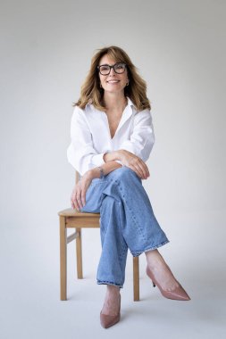 Sarışın kadın izole edilmiş arka planda güvenle oturuyor ve kameraya bakıyor. Beyaz gömlek ve kot pantolon giyen orta yaşlı bir kadın. Uzayı kopyala Tam uzunluk.