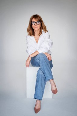 Sarışın kadın izole edilmiş arka planda güvenle oturuyor ve kameraya bakıyor. Beyaz gömlek ve kot pantolon giyen orta yaşlı bir kadın. Uzayı kopyala Tam uzunluk.
