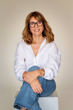 Sarışın kadın izole edilmiş arka planda güvenle oturuyor ve kameraya bakıyor. Beyaz gömlek ve kot pantolon giyen orta yaşlı bir kadın. Boşluğu kopyala. 
