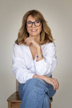 Sarışın kadın izole edilmiş arka planda güvenle oturuyor ve kameraya bakıyor. Beyaz gömlek ve kot pantolon giyen orta yaşlı bir kadın. Boşluğu kopyala. 