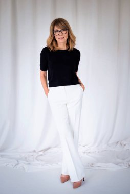 Gülümseyen olgun bir kadının portresi. Kendine güvenen, sarı saçlı, açık tenli bir kadın. Siyah kazak ve beyaz pantolon giyen çekici bir kadın. Uzayı kopyala Tam uzunluk.