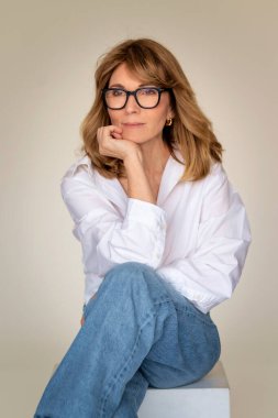 Sarışın kadın özgüvenle arka planda oturuyor ve özenle bakıyor. Beyaz gömlek ve kot pantolon giyen orta yaşlı bir kadın. Boşluğu kopyala. 