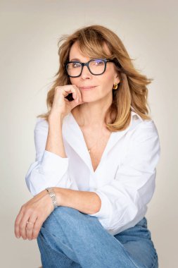 Sarışın kadın izole edilmiş arka planda güvenle oturuyor ve kameraya bakıyor. Beyaz gömlek ve kot pantolon giyen orta yaşlı bir kadın. Boşluğu kopyala. 