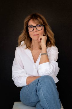 Sarışın kadın, izole edilmiş siyah arka planda güvenle oturuyor ve kameraya bakıyor. Beyaz gömlek ve kot pantolon giyen orta yaşlı bir kadın. Boşluğu kopyala. 