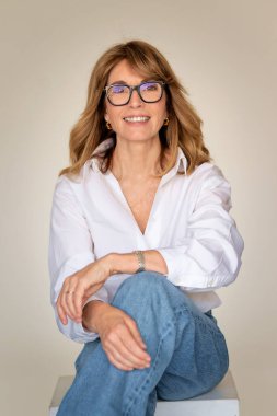 Sarışın kadın izole edilmiş arka planda güvenle oturuyor ve kameraya bakıyor. Beyaz gömlek ve kot pantolon giyen orta yaşlı bir kadın. Boşluğu kopyala. 