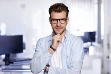 Modern bir ofiste tek başına dururken gülümseyen kendinden emin genç bir iş adamının portresi. Gündelik kıyafetler giyen genç bir girişimci. Yakın plan çekim. Eller çenede..