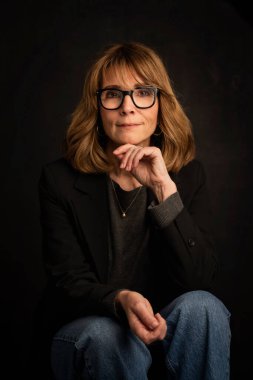 Sarışın kadın, izole edilmiş siyah arka planda güvenle oturuyor ve kameraya bakıyor. Ceket ve kot pantolon giyen orta yaşlı bir kadın. Boşluğu kopyala. 