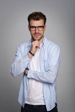 Tecrit edilmiş arka planda duran yakışıklı bir delikanlının yakın çekimi. Uzayı kopyala Tişört ve gözlük takan kendinden emin bir erkek. Eller çenede..