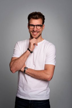 Tecrit edilmiş arka planda duran yakışıklı bir delikanlının yakın çekimi. Uzayı kopyala Beyaz tişört ve gözlük takan kendinden emin bir erkek. Eller çenede..