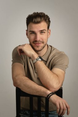 Tecrit edilmiş arka planda oturan yakışıklı bir gencin stüdyo fotoğrafı. Polo tişört giyen ve kameraya bakan kendinden emin bir erkek. Uzayı kopyala İzole edilmiş arkaplan.