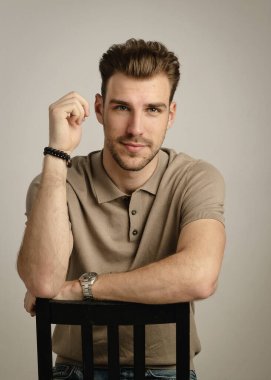 Tecrit edilmiş arka planda oturan yakışıklı bir gencin stüdyo fotoğrafı. Polo tişört giyen ve kameraya bakan kendinden emin bir erkek. Uzayı kopyala İzole edilmiş arkaplan.