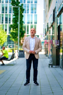 Şehir caddesinde duran 50 'li yaşlarda kendine güvenen bir adam. Ceket ve siyah pantolon giyen kel ve sakallı bir erkek. - Evet. Modern ofis binaları ve arka planda yeşil konuşmalar. Tam boy çekim. 