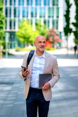 Yakışıklı bir adam dizüstü bilgisayar tutuyor ve şehirde finans sektöründe yürüyor. Orta yaşlı iş adamı günlük iş kıyafetleri ve kısa mesajlar giyiyor.. 