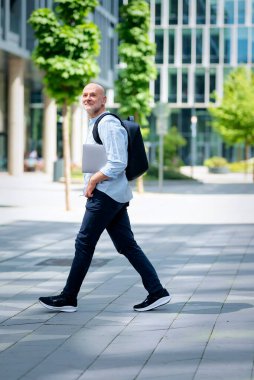 50 'li yaşlarda şehir caddesinde yürüyen kendine güvenen bir adam. Kel ve sakallı erkek, gömlek ve siyah pantolon giyiyor. Omzunda siyah bir sırt çantası ve elinde bir dizüstü bilgisayar vardı. Modern ofis binaları ve arka planda yeşil konuşmalar.
