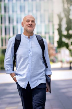 50 'li yaşlarda şehir caddesinde yürüyen kendine güvenen bir adam. Kel ve sakallı erkek, gömlek ve siyah pantolon giyiyor. Omzunda siyah bir sırt çantası ve elinde bir dizüstü bilgisayar vardı. Modern ofis binaları ve arka planda yeşil konuşmalar.