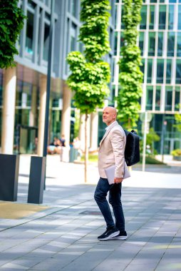 50 'li yaşlarda şehir caddesinde yürüyen kendine güvenen bir adam. Ceket ve siyah pantolon giyen kel ve sakallı bir erkek. Omzunda siyah bir sırt çantası ve elinde bir dizüstü bilgisayar vardı. Modern ofis binaları ve arka planda yeşil konuşmalar.
