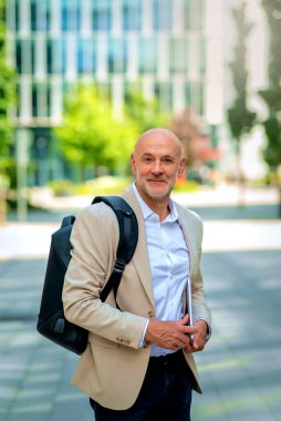 50 'li yaşlarda şehir caddesinde yürüyen kendine güvenen bir adam. Ceket ve siyah pantolon giyen kel ve sakallı bir erkek. Omzunda siyah bir sırt çantası ve elinde bir dizüstü bilgisayar vardı. Modern ofis binaları ve arka planda yeşil konuşmalar.