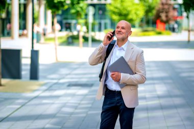 Yakışıklı bir adam dizüstü bilgisayar tutuyor ve şehirde finans sektöründe yürüyor. Orta yaşlı işadamı iş kıyafeti giyiyor ve telefon görüşmesi yapıyor.. 
