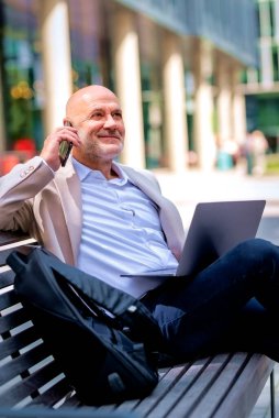 Profesyonel erkek, dışarıda bir bankta oturuyor, dizüstü bilgisayar kullanırken bir telefon görüşmesi yapıyor. Uzaktan çalışan olgun bir adam, cep telefonu ve bilgisayar laptopuyla birden fazla iş yapıyor..