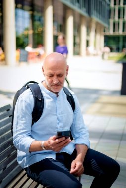 50 'li yaşlarda bir bankta oturan ve elinde akıllı telefon tutan kendinden emin bir adam. Kel ve sakallı erkek, gömlek ve siyah pantolon giyiyor. Omzunda siyah bir sırt çantası vardı. Modern ofis binaları ve arka planda yeşil konuşmalar.