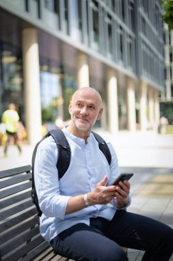 50 'li yaşlarda bir bankta oturan ve elinde akıllı telefon tutan kendinden emin bir adam. Kel ve sakallı erkek, gömlek ve siyah pantolon giyiyor. Omzunda siyah bir sırt çantası vardı. Modern ofis binaları ve arka planda yeşil konuşmalar.
