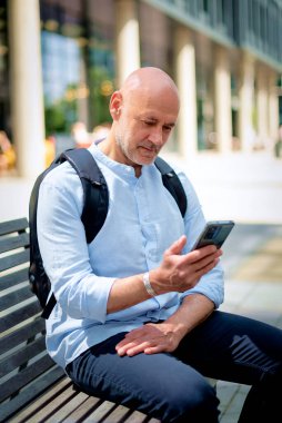50 'li yaşlarda bir bankta oturan ve elinde akıllı telefon tutan kendinden emin bir adam. Kel ve sakallı erkek, gömlek ve siyah pantolon giyiyor. Omzunda siyah bir sırt çantası vardı. Modern ofis binaları ve arka planda yeşil konuşmalar.