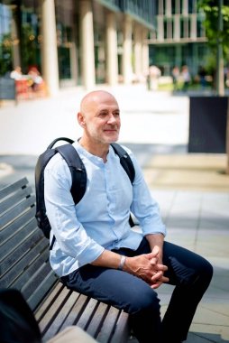 50 'li yaşlarda kenar mahallede bankta oturan kendinden emin bir adam. Kel ve sakallı erkek, gömlek ve siyah pantolon giyiyor. Omzunda siyah bir sırt çantası vardı. Modern ofis binaları ve arka planda yeşil konuşmalar.