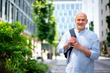 50 'li yaşlarda şehir caddesinde yürüyen kendine güvenen bir adam. Kel ve sakallı erkek, gömlek ve siyah pantolon giyiyor. Omzunda siyah bir sırt çantası vardı. Modern ofis binaları ve arka planda yeşil konuşmalar.